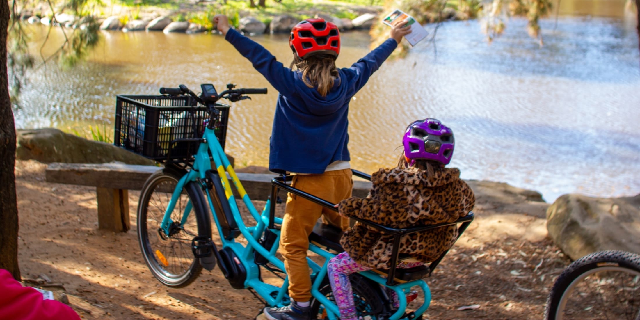 Two children in helmets beside a river with an aqua Tribe Evamos Longtail, one standing on the rear rack with both arms raised