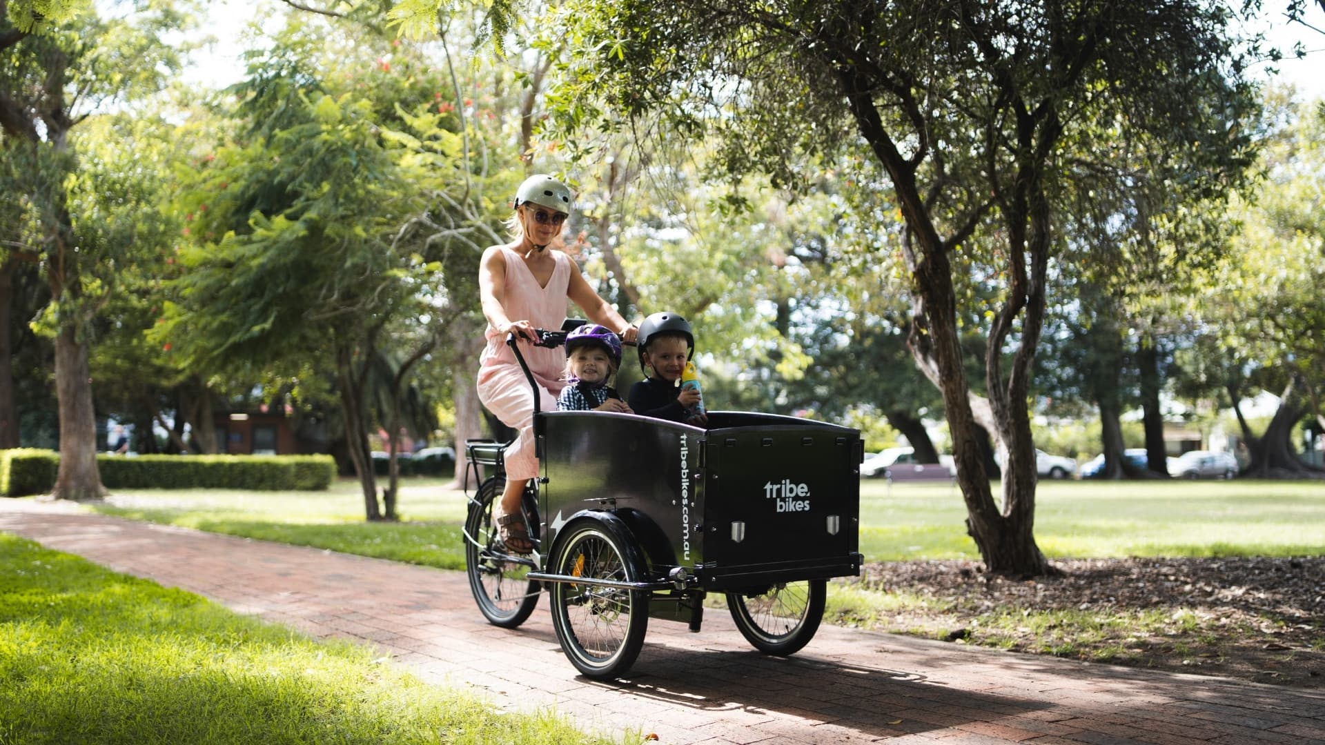 A mum in a white helmet riding a black Tribe Original along a dappled brick park path, two small children in helmets sitting in the front cargo box, one holding a drink bottle