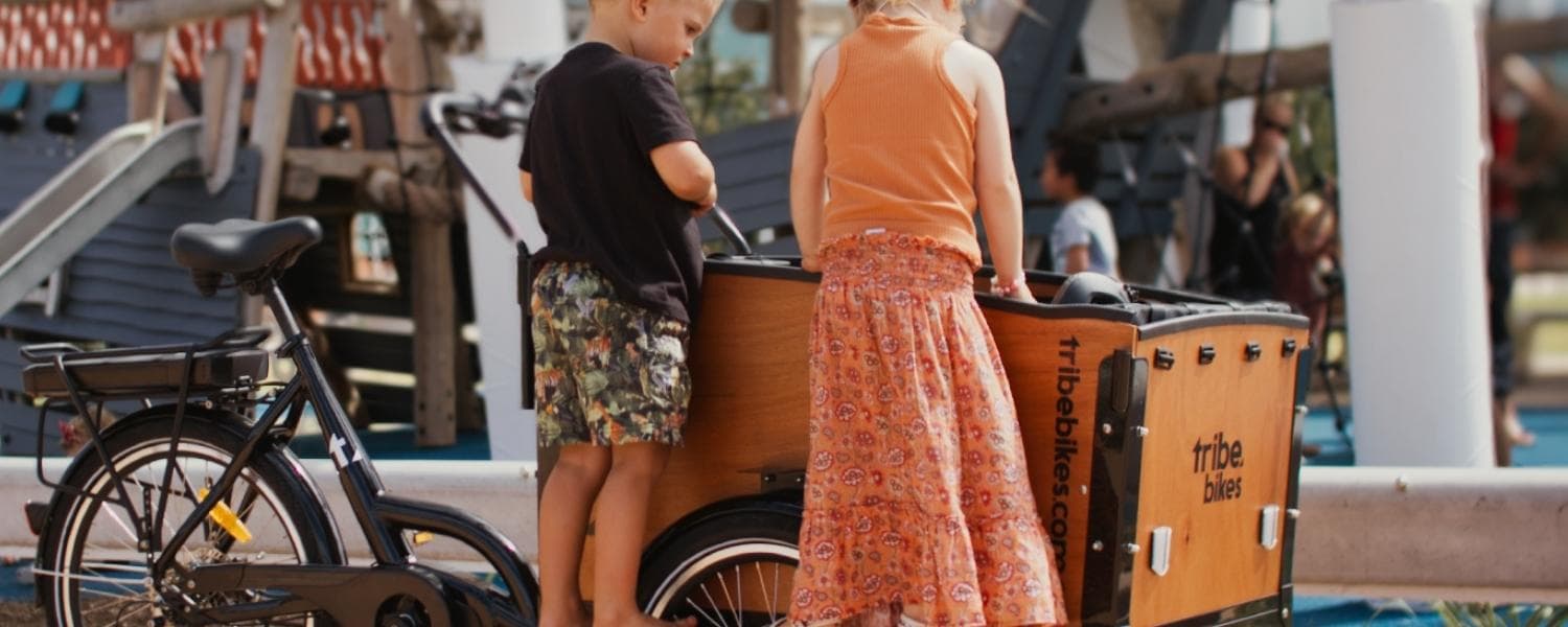 Two young children at a playground, backs to the camera, leaning over the open plywood cargo box of a Tribe Original