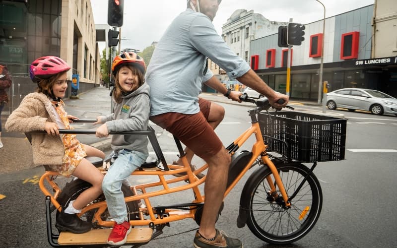 A dad riding an orange Tribe Evamos Longtail across a city intersection with two helmeted children on the rear seat and a crate on the front rack