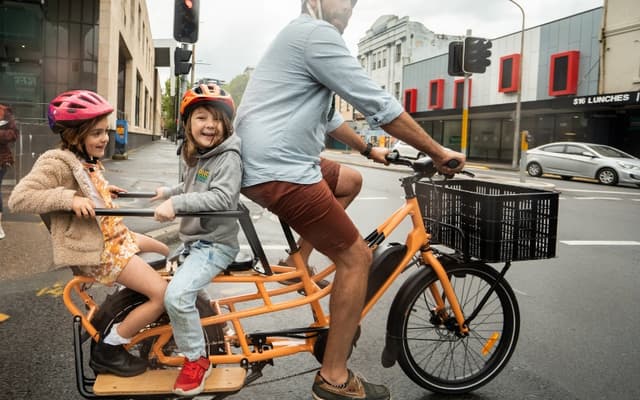 A dad riding an orange Tribe Evamos Longtail across a city intersection with two helmeted children on the rear seat and a crate on the front rack