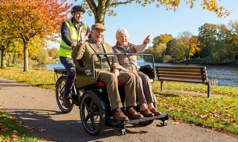 Two older passengers waving from a Ricky Rickshaw on a riverside path in autumn, with a volunteer pilot riding behind